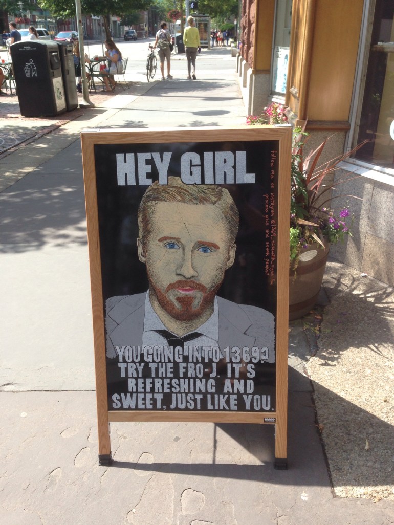A picture of a sandwich board in front of a cafe, with an illustration of Ryan Seacrest, that says 'Hey Girl, you going into 1369? Try the Fro-J. It's refreshing and sweet, just like you.'